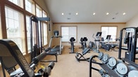 Located at 3025 Fountain Dr. in Beavercreek, Ohio, this single-floor fitness center is a comprehensively outfitted space designed to support a full range of workout routines. The room is defined by soaring vaulted ceilings accented by a ceiling fan and recessed lighting, creating an open and airy atmosphere. Neutral-toned walls and warm wood trim details lend the space a clean, welcoming aesthetic, while carpeted flooring provides comfort underfoot throughout the facility.

Natural light flows generously through multiple windows fitted with blinds, keeping the environment bright and inviting. The cardio section includes treadmills, upright and recumbent bikes, and an elliptical machine, offering a variety of options for cardiovascular training. Strength training is equally well-represented, with a cable crossover machine, a Nautilus multi-station unit, a power rack with an adjustable bench, and a dedicated dumbbell rack stocked with a full range of free weights and weight plates.

Additional equipment such as a stability ball, a wall-mounted flat-screen television, a large mirror, and a coat hook station further enhance the functionality and convenience of the space. French doors provide access to the fitness area, adding an architectural touch that complements the overall design. This facility is thoughtfully arranged to accommodate both individual training sessions and group use with ease.