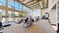 This fitness center offers a generously sized, single-floor workout facility designed to provide a premium exercise experience. The space is defined by its dramatic double-height ceilings adorned with decorative coffered beams and stylish pendant light fixtures, creating an open and airy atmosphere throughout.

Floor-to-ceiling windows line the perimeter, flooding the interior with abundant natural light and offering expansive views of the surrounding outdoor landscape and streetscape. The patterned carpet flooring adds a refined, upscale aesthetic that distinguishes this facility from a standard gym environment.

The equipment layout is thoughtfully organized to maximize usability. A dedicated cardio zone features multiple treadmills and elliptical trainers, including TechnoGym brand machines, all positioned along the windowed wall to allow users to enjoy the outdoor views during workouts. A strength training section houses a wide array of selectorized cable and resistance machines arranged in rows throughout the main floor area.

A separate free weights area includes a full dumbbell rack, flat benches, a Smith machine, medicine balls, and additional specialty equipment, complemented by a large wall mirror and a wall-mounted flat-screen television. Recessed lighting, alongside the pendant fixtures, ensures consistent illumination across all zones. The overall design balances functionality with an upscale aesthetic, making it a well-rounded and inviting fitness destination.