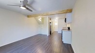 This two-floor space showcases a thoughtfully designed open-plan layout that seamlessly connects the living area to the kitchen. A standout architectural feature is the exposed natural wood beam that spans the ceiling, adding rustic warmth and visual interest to the otherwise clean, modern interior. Dark hardwood flooring runs throughout, providing a rich contrast to the bright white walls and creating a cohesive flow across the space.

The living area is generously sized, with a ceiling fan providing both functionality and a casual aesthetic. A small pendant light cluster adds a decorative touch, illuminating the transition zone between the living and kitchen areas. The angled ceiling near the stairwell hints at the second floor above, adding architectural dimension.

The kitchen is compact yet well-appointed, featuring white cabinetry, granite-look countertops, stainless steel appliances including a refrigerator and range, and an under-cabinet range hood. A small window near the rear of the unit allows natural light to filter into the space. Two doors at the back of the unit suggest access to additional rooms or storage, contributing to the overall functional layout of this multi-level space.