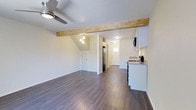 This two-floor space showcases a thoughtfully designed open-plan layout that seamlessly connects the living area to the kitchen. A standout architectural feature is the exposed natural wood beam that spans the ceiling, adding rustic warmth and visual interest to the otherwise clean, modern interior. Dark hardwood flooring runs throughout, providing a rich contrast to the bright white walls and creating a cohesive flow across the space.

The living area is generously sized, with a ceiling fan providing both functionality and a casual aesthetic. A small pendant light cluster adds a decorative touch, illuminating the transition zone between the living and kitchen areas. The angled ceiling near the stairwell hints at the second floor above, adding architectural dimension.

The kitchen is compact yet well-appointed, featuring white cabinetry, granite-look countertops, stainless steel appliances including a refrigerator and range, and an under-cabinet range hood. A small window near the rear of the unit allows natural light to filter into the space. Two doors at the back of the unit suggest access to additional rooms or storage, contributing to the overall functional layout of this multi-level space.