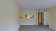 This single-story space features a straightforward, functional layout with a seamless connection between the living area and kitchen. The living room is finished with wall-to-wall neutral-toned carpeting, providing a comfortable and cohesive look throughout. Walls are painted in a soft, off-white tone, contributing to a clean and bright atmosphere.

A defining architectural feature is the pass-through cutout in the shared wall between the living area and kitchen, allowing for an open sightline and improved interaction between the two spaces. The kitchen, visible through this opening, is equipped with standard appliances including a stovetop and overhead microwave, with white cabinetry and under-cabinet lighting offering a practical and tidy workspace.

Ceiling-mounted HVAC vents are positioned throughout, ensuring efficient climate control across the space. A doorway off the main living area leads to an additional room, likely a hallway or bathroom, extending the overall functionality of the unit. The overall design is modest and utilitarian, emphasizing ease of use and straightforward livability in a compact, single-level layout.