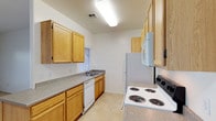 The kitchen is designed with functionality in mind, featuring a galley-style layout that maximizes efficiency within the available space. Warm honey-toned wood cabinetry lines both walls, providing generous storage above and below the countertops. The speckled grey laminate countertops offer a practical work surface throughout. White appliances, including a coil-burner electric range, built-in over-the-range microwave, refrigerator, and dishwasher, are neatly integrated into the layout. A double-basin sink is positioned beneath a window, allowing natural light to brighten the workspace. Light-toned tile flooring runs throughout, complementing the neutral white walls. Overhead fluorescent lighting ensures the kitchen remains well-lit. The clean, straightforward design prioritizes practicality, making the most of the corridor layout with thoughtful placement of appliances and cabinetry to create an organized and efficient cooking environment.