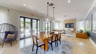 Located at 9051 SW 122nd Ave in Miami, FL, this single-story community clubhouse delivers a polished, resort-inspired atmosphere with thoughtfully curated interiors throughout. The grand entrance lobby immediately sets the tone with soaring double-height ceilings, gleaming travertine tile flooring, and a striking vertical slat wall installation that draws the eye upward. A bold living moss wall accent flanks the leasing office area, where a sleek workstation is paired with warm yellow accent chairs and a built-in wood shelving unit displaying curated décor and accessories. Adjacent to the lobby, a welcoming lounge features a tufted charcoal leather Chesterfield sofa, textured area rugs, and abundant natural light streaming through oversized windows with lush outdoor views. The dining area is a centerpiece of the space, anchored by a substantial solid-wood live-edge dining table beneath globe pendant lights, complemented by a wet bar with bold blue tile accents. A separate lounge nook offers a cognac leather sectional set against a wood-paneled TV wall with cool blue accent walls and modern artwork. A curated gallery wall near the entry, styled with eclectic framed art and a woven-front credenza, adds a welcoming, residential warmth to this vibrant community gathering space.