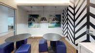 This coffee bar is a thoughtfully designed communal space that balances vibrant aesthetics with practical functionality. The most striking visual element is a bold black-and-white chevron/herringbone graphic accent wall, which anchors the beverage station and immediately establishes the space's modern, energetic character.

The beverage station features a professional-grade coffee machine, white cabinetry with sleek hardware, and a marble-style countertop, providing a well-equipped self-serve setup. Light wood-look flooring runs throughout, lending warmth and continuity across the different seating zones.

The seating areas are diverse and flexible. Round metallic tables paired with bold cobalt blue upholstered ottomans create casual lounge clusters, while black square tables with contemporary chairs offer more structured seating. Overhead, geometric pendant lights hang above the lounge area, complementing a pair of abstract canvas artworks in teal and green tones that add an artistic focal point.

Glass-partitioned walls and sliding doors connect the coffee bar to adjacent meeting rooms, allowing natural visual flow and light between spaces while maintaining acoustic separation. The overall ambiance is lively yet refined, combining bold design choices with a welcoming, collaborative atmosphere suited for both breaks and informal work sessions.