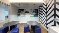 This coffee bar is a thoughtfully designed communal space that balances vibrant aesthetics with practical functionality. The most striking visual element is a bold black-and-white chevron/herringbone graphic accent wall, which anchors the beverage station and immediately establishes the space's modern, energetic character.

The beverage station features a professional-grade coffee machine, white cabinetry with sleek hardware, and a marble-style countertop, providing a well-equipped self-serve setup. Light wood-look flooring runs throughout, lending warmth and continuity across the different seating zones.

The seating areas are diverse and flexible. Round metallic tables paired with bold cobalt blue upholstered ottomans create casual lounge clusters, while black square tables with contemporary chairs offer more structured seating. Overhead, geometric pendant lights hang above the lounge area, complementing a pair of abstract canvas artworks in teal and green tones that add an artistic focal point.

Glass-partitioned walls and sliding doors connect the coffee bar to adjacent meeting rooms, allowing natural visual flow and light between spaces while maintaining acoustic separation. The overall ambiance is lively yet refined, combining bold design choices with a welcoming, collaborative atmosphere suited for both breaks and informal work sessions.