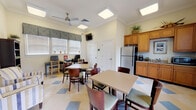 This single-floor community space is thoughtfully designed to serve as a comfortable and functional gathering area for its residents. The main room features a distinctive checkered tile floor in cream and blue tones, lending a clean and cheerful aesthetic throughout. Multiple dining tables with padded chairs are arranged to accommodate group meals or social gatherings, while a ceiling fan with flush-mounted panel lighting keeps the space bright and well-ventilated.

A dedicated kitchenette area is outfitted with warm honey-toned wood cabinetry, granite-style countertops, a stainless steel refrigerator, a double sink, a microwave, and a coffee maker — providing convenient access to refreshments and light meal preparation. Natural light filters through white-shuttered windows, enhancing the inviting atmosphere.

A cozy lounge nook features a pair of striped upholstered armchairs positioned beside a tall bookcase stocked with reading materials, alongside framed artwork that adds a decorative touch to the cream-colored walls. Soaring ceilings with crown molding lend the space an open, airy feel. A computer workstation area with monitors and chairs is also available, and a wall-mounted television provides additional entertainment. French glass-paned doors connect to adjacent areas, contributing to a bright, open layout throughout.