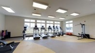 This fitness center offers a bright, welcoming environment designed to support a range of workout routines. The space is defined by a row of large, white-framed windows that span the length of one wall, flooding the interior with natural light and offering open outdoor views — creating an airy, motivating atmosphere during workouts. Overhead, a series of recessed panel light fixtures supplement the natural light, ensuring the space is well-illuminated at all times. The flooring features a durable, multi-tone exercise surface with curved zoning, providing both functionality and visual interest. The cardio equipment lineup includes multiple treadmills and elliptical trainers, each equipped with individual display screens, as well as a stationary bike, catering to a variety of fitness preferences. Stability balls are available along the perimeter for stretching and core training exercises. The neutral color palette of the walls and ceiling contributes to a clean, focused environment. The overall layout is open and uncluttered, allowing for ease of movement between stations. This fitness center is thoughtfully designed to provide members with a comfortable and fully functional space for their daily exercise needs.