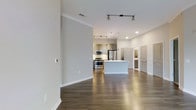This single-floor space showcases a contemporary open-concept design that seamlessly connects the living area to the kitchen. The rich, dark hardwood flooring runs throughout, providing a warm contrast to the crisp white walls and bright, airy atmosphere created by the recessed and track lighting overhead.

The kitchen is a standout feature, equipped with stainless steel appliances including a range, microwave, and refrigerator. A central island with a clean white finish serves as both a functional workspace and a natural focal point of the room. The cabinetry features a modern taupe-toned finish, complementing the neutral color palette of the space.

Crown molding adds a refined architectural detail along the upper walls, enhancing the overall polished aesthetic. The open layout creates a generous sense of space, allowing for flexible furniture arrangements in the living area.

Toward the rear of the space, multiple doorways lead to additional rooms, with interior doors featuring a raised-panel design in a soft grey finish. The overall ambiance is clean, modern, and well-lit, making it a highly functional and visually cohesive living environment.