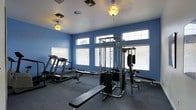 This single-floor fitness space is designed to support a well-rounded workout routine. The room is finished with bold blue walls that create an energetic atmosphere, complemented by a neutral gray carpet underfoot. Natural light filters in through multiple windows positioned along the upper portion of the walls, fitted with white horizontal blinds that allow for light control while maintaining privacy. The ceiling is white and features warm-toned globe light fixtures that provide supplemental illumination throughout the space. Equipment includes a multi-station cable weight machine positioned centrally, offering a variety of strength-training exercises, alongside a flat weight bench for free-weight workouts. A treadmill occupies one corner of the room, providing a cardio option. The layout is functional and organized, with equipment spaced to allow for comfortable use. Ventilation is supported by ceiling vents integrated into the white ceiling. Overall, the space offers a practical and focused environment for fitness activities, balancing natural brightness with dedicated workout equipment.