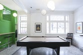 This single-floor communal space masterfully blends classic architectural details with contemporary design sensibilities. The expansive layout is anchored by striking polychrome terrazzo floors featuring bold stripe inlays that guide residents through distinct functional zones. White-painted walls, prominent crown molding, and tall multi-pane windows flood the interior with natural light while reinforcing an elegant, timeless character.

A dramatic focal point is the vivid green staircase structure, boldly painted and outfitted with bar-height seating beneath, creating a casual gathering spot at the heart of the space. Flanking the staircase, sturdy white columns frame an open great room complete with a full-size billiards table, making it ideal for recreation and socializing.

The lounge area features a vibrant orange sectional sofa paired with a sculptural white accent chair and a patterned area rug, offering a warm and inviting retreat. Nearby, a low-profile platform seating unit with plywood cabinetry faces a wall-mounted television surrounded by a thoughtfully curated gallery of framed artwork. Spherical pendant lights provide warm, ambient illumination throughout.

A dedicated mailroom with individual resident mailboxes and an iconic Egg Chair provides a functional yet stylish entry point. The overall ambiance is creative, contemporary, and community-focused.