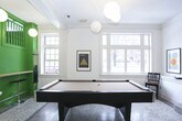 This single-floor communal space masterfully blends classic architectural details with contemporary design sensibilities. The expansive layout is anchored by striking polychrome terrazzo floors featuring bold stripe inlays that guide residents through distinct functional zones. White-painted walls, prominent crown molding, and tall multi-pane windows flood the interior with natural light while reinforcing an elegant, timeless character.

A dramatic focal point is the vivid green staircase structure, boldly painted and outfitted with bar-height seating beneath, creating a casual gathering spot at the heart of the space. Flanking the staircase, sturdy white columns frame an open great room complete with a full-size billiards table, making it ideal for recreation and socializing.

The lounge area features a vibrant orange sectional sofa paired with a sculptural white accent chair and a patterned area rug, offering a warm and inviting retreat. Nearby, a low-profile platform seating unit with plywood cabinetry faces a wall-mounted television surrounded by a thoughtfully curated gallery of framed artwork. Spherical pendant lights provide warm, ambient illumination throughout.

A dedicated mailroom with individual resident mailboxes and an iconic Egg Chair provides a functional yet stylish entry point. The overall ambiance is creative, contemporary, and community-focused.
