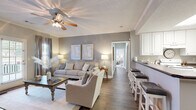 This single-floor residence offers a thoughtfully designed layout that balances comfort and functionality throughout. The living room serves as the heart of the home, featuring warm hardwood-style flooring, crown molding, a ceiling fan, and neutral gray walls that create a calm, welcoming ambiance. A glass door floods the space with natural light and provides access to the outdoors. Adjacent to the living area, a peninsula-style kitchen showcases white cabinetry, dark countertops, and a breakfast bar with seating — seamlessly connecting to a dedicated dining room adorned with crown molding and a pendant light overhead. The bedrooms are carpeted and generously sized, each equipped with a ceiling fan and windows that invite natural light. One bedroom includes a spacious walk-in closet outfitted with dual hanging rods and built-in shelving. The primary bathroom features a dual-sink vanity with white cabinetry, brushed nickel fixtures, and a large mirror, conveying a clean and polished aesthetic. A dedicated laundry room with full-size washer and dryer hookups and overhead cabinetry adds everyday convenience. Throughout the home, a cohesive neutral color scheme, consistent trim work, and tasteful finishes contribute to a refined yet comfortable living environment.