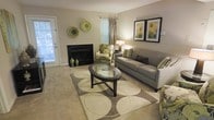 This single-floor space centers around a well-appointed living room designed with a cohesive neutral color palette of warm taupes and greiges, accented by touches of green throughout. The focal point of the room is a sleek, black fireplace framed by light-colored walls, adding both warmth and character to the space. An oval glass-top coffee table with a dark wood base anchors the seating area, which features a plush grey sofa dressed with green and grey accent pillows. A floral-patterned armchair in coordinating green tones adds visual interest and complements the room's color scheme. Decorative woven wall medallions in earthy greens are displayed above the fireplace, while a pair of framed abstract artworks adds a polished finishing touch to the opposite wall. A patterned area rug with large circular motifs in cream and sage green defines the seating area over the neutral carpeted floor. Natural light filters in through a draped window and a glass-paneled exterior door, keeping the space bright and airy. The overall ambiance is comfortable and tastefully styled, balancing casual living with refined décor details.