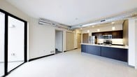 This single-floor unit showcases an open-concept layout that seamlessly integrates the living area and kitchen. A defining industrial design element is the exposed metallic ductwork running along the ceiling, adding an urban loft character to the space. The ceiling is otherwise clean and white, contributing to a bright and airy atmosphere.

The kitchen is positioned along one side of the space and features a central island with a light countertop and sleek navy-blue cabinetry below. Dark upper cabinetry lines the back wall alongside stainless steel appliances, including a refrigerator, microwave, and range. Pendant lighting above the island provides focused task illumination while enhancing the kitchen's contemporary style.

The living area is open and generously proportioned, finished with light-toned flooring that reflects available light and creates a sense of spaciousness. Neutral white walls maintain a clean backdrop throughout. Built-in closets and a doorway visible near the rear of the space suggest additional storage and room access. A floor vent and wall outlets indicate practical utility throughout. Overall, the space balances modern industrial design with functional everyday living.