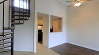 This two-story space makes an immediate impression with its dramatically tall ceilings in the main living area, creating a sense of openness and volume. A ceiling fan is mounted at height, helping to circulate air throughout the expansive vertical space. The living area features a combination of flooring materials, with hardwood-style planks transitioning to plush carpeting, defining separate zones within the open layout.

A staircase with a dark metal railing is visible along one wall, leading to the upper level and adding a bold architectural contrast against the clean white walls. The kitchen is accessible through a dedicated doorway and is equipped with dark appliances, including a black range, complemented by light wood cabinetry. A pass-through window connects the kitchen to an adjacent area, enhancing sightlines and communication between spaces.

The overall aesthetic is clean and neutral, with white walls throughout providing a bright, versatile backdrop. The layout reflects a practical, well-organized design, with clearly defined areas that flow naturally from one to the next across both floors.