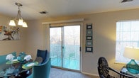 This single-floor residence offers a welcoming and functional layout designed for comfortable everyday living. The living and dining area serves as the heart of the home, featuring plush carpeting underfoot and neutral-toned walls that create a warm, versatile backdrop. A standout feature of this space is the large sliding glass door that floods the interior with natural light and opens directly onto a private balcony, where outdoor seating invites relaxation and enjoyment of the surrounding tree-lined views. The balcony is enclosed with a decorative lattice railing, adding a charming touch while maintaining an open, airy feel. The dining area is furnished with a round table and vibrant teal upholstered chairs, adding a lively pop of color to the room. Decorative wall art displayed near the balcony door adds a personalized, curated touch to the space. A ceiling-mounted HVAC vent ensures year-round climate comfort throughout the interior. The overall ambiance is bright, casual, and inviting, making it a pleasant and practical living environment suitable for a variety of lifestyles.