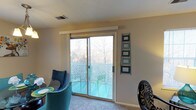 This single-floor residence offers a welcoming and functional layout designed for comfortable everyday living. The living and dining area serves as the heart of the home, featuring plush carpeting underfoot and neutral-toned walls that create a warm, versatile backdrop. A standout feature of this space is the large sliding glass door that floods the interior with natural light and opens directly onto a private balcony, where outdoor seating invites relaxation and enjoyment of the surrounding tree-lined views. The balcony is enclosed with a decorative lattice railing, adding a charming touch while maintaining an open, airy feel. The dining area is furnished with a round table and vibrant teal upholstered chairs, adding a lively pop of color to the room. Decorative wall art displayed near the balcony door adds a personalized, curated touch to the space. A ceiling-mounted HVAC vent ensures year-round climate comfort throughout the interior. The overall ambiance is bright, casual, and inviting, making it a pleasant and practical living environment suitable for a variety of lifestyles.