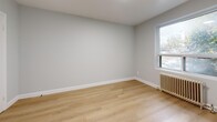 This thoughtfully designed 1-bedroom, 1-bathroom unit is laid out across a single floor, encompassing a living room, bedroom, kitchen, hallway, and bathroom. Throughout the unit, light-toned hardwood flooring provides a warm, cohesive aesthetic that complements the consistently neutral, soft-gray painted walls and clean white baseboards.

The living room offers an open and airy feel with recessed lighting and a window framing views of mature surrounding trees, allowing natural light to filter in. A classic cast-iron radiator beneath the window serves as both a functional and character-rich detail.

The bedroom is a comfortable, well-proportioned space featuring its own window with a radiator, recessed lighting, and two interior doors — one leading to the hallway and another providing additional access or closet space. The room maintains the same clean, minimalist aesthetic found throughout the unit.

The kitchen is equipped with white shaker-style cabinetry, granite-look countertops, and a stainless steel refrigerator, creating a modern and functional cooking space. The bathroom features a full soaking tub with crisp white tile surround, offering a clean and serene finish.

A connecting hallway ties all rooms together efficiently, completing this compact yet well-organized home.