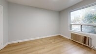 This thoughtfully designed 1-bedroom, 1-bathroom unit is laid out across a single floor, encompassing a living room, bedroom, kitchen, hallway, and bathroom. Throughout the unit, light-toned hardwood flooring provides a warm, cohesive aesthetic that complements the consistently neutral, soft-gray painted walls and clean white baseboards.

The living room offers an open and airy feel with recessed lighting and a window framing views of mature surrounding trees, allowing natural light to filter in. A classic cast-iron radiator beneath the window serves as both a functional and character-rich detail.

The bedroom is a comfortable, well-proportioned space featuring its own window with a radiator, recessed lighting, and two interior doors — one leading to the hallway and another providing additional access or closet space. The room maintains the same clean, minimalist aesthetic found throughout the unit.

The kitchen is equipped with white shaker-style cabinetry, granite-look countertops, and a stainless steel refrigerator, creating a modern and functional cooking space. The bathroom features a full soaking tub with crisp white tile surround, offering a clean and serene finish.

A connecting hallway ties all rooms together efficiently, completing this compact yet well-organized home.