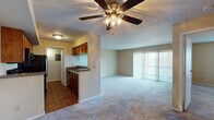This single-floor residence is thoughtfully laid out to maximize functionality and comfort. The open-concept living area is spacious and carpeted, anchored by a ceiling fan with integrated lighting and a textured ceiling. A set of sliding glass doors with vertical blinds leads to a private balcony, allowing natural light to fill the room and providing outdoor views.

The kitchen connects seamlessly to the living space via a pass-through opening, creating a cohesive and social floor plan. It features warm-toned wood cabinetry, granite-style countertops, and a full suite of black appliances including a refrigerator, electric range, over-the-range microwave, and dishwasher. Hardwood-style flooring runs throughout the kitchen and utility areas, adding a practical contrast to the carpeted living spaces.

Two well-proportioned bedrooms each feature plush carpeting, neutral wall tones, and windows fitted with blinds that allow for natural light and privacy. The primary bedroom benefits from two windows and is situated near one of the two full bathrooms.

Both bathrooms include a tub-shower combination with white surround panels, a vanity with wood cabinetry, and hardwood-style flooring. The layout as a whole offers a practical and comfortable living environment with clearly defined zones for cooking, relaxing, sleeping, and bathing.