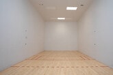 This single-level space is purpose-built as a racquetball court, designed to meet the functional demands of both recreational and competitive players. The court is defined by its polished, light-toned hardwood flooring, finished to a smooth surface that ensures consistent ball bounce and safe footing. Red court lines are precisely marked across the floor, clearly delineating the service zones and boundary areas required for regulation play.

One of the most notable architectural features of this court is its impressively tall ceiling height, which provides ample vertical clearance essential for racquetball gameplay. The walls are finished in clean, bright white, maximizing light reflection and ensuring excellent visibility throughout the playing area. Overhead, recessed flush-mounted skylight panels supply even, diffused artificial lighting across the entire court surface.

A full-height glass wall and glass door entry system occupies one end of the court, offering a transparent partition that allows spectators to observe play from outside without obstruction. Through the glass, an adjacent fitness area is visible, suggesting this court is part of a broader recreational facility. A small informational placard is mounted on the interior wall near the entry, and a waste bin is conveniently positioned near the door. The overall environment is clean, minimalist, and well-maintained.