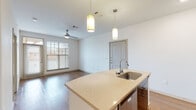This well-designed single-floor residence offers a clean, contemporary aesthetic throughout its open-concept layout. The kitchen serves as the centerpiece of the main living area, anchored by a spacious island with a light stone countertop, an undermount stainless steel sink, and elegant dual pendant lights hanging overhead. Dark espresso cabinetry lines the kitchen walls, complemented by a tiled backsplash and a full suite of stainless steel appliances, including a range, built-in microwave, dishwasher, and refrigerator. The kitchen flows seamlessly into the living area, which features warm wood-look flooring, crisp white walls, and a ceiling fan. Natural light filters in through a large window fitted with horizontal blinds, brightening the open space. A bedroom off the main living area is finished with plush carpeting, its own ceiling fan, and a window that invites additional natural light. The bathroom is efficiently laid out with matching stone countertops and wood-look flooring consistent with the rest of the unit. Double doors near the entryway provide generous storage or closet space. The overall ambiance is bright, modern, and move-in ready, with thoughtful finishes carried consistently throughout.