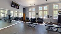 Located at 101 Legacy Terrace in Chapel Hill, NC, this fitness center is a well-appointed, single-floor facility designed to support a comprehensive workout experience. The space is bathed in natural light, thanks to large, multi-pane windows that line the walls and offer views of the surrounding greenery, creating an open and energizing atmosphere. The interior features soft blue-gray walls, durable rubber flooring, and elevated ceilings with overhead lighting and ceiling fans that promote airflow and comfort throughout. The cardio zone is equipped with multiple treadmills and elliptical machines, each positioned to take advantage of the natural light and exterior views. Flat-screen televisions are mounted throughout the space for entertainment during workouts. A dedicated strength training area includes a multi-functional cable machine, a power rack, weight benches, and an extensive dumbbell rack with a full-length mirror for form checks. Additional functional training equipment such as TRX suspension trainers and a heavy punching bag add versatility to the space. The facility also features direct exterior access through glass-paneled doors, connecting the interior to an outdoor area. Overall, the fitness center provides a spacious, well-lit, and thoroughly equipped environment suited for a wide range of fitness routines.