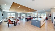 Located in Summerfield, Florida, this generously sized single-floor community room is a well-appointed gathering space designed for socializing and recreation. The open-plan layout is anchored by a striking central island with a crisp white countertop and slate-blue cabinetry, accompanied by bar-height seating. The full kitchen behind it features white shaker-style cabinetry with glass-front uppers, a bold vertical dark tile backsplash, stainless steel appliances, and stylish cone-shaped pendant lighting. A signature tray ceiling with a warm gold finish adds architectural depth to the main gathering area, which is illuminated by recessed lighting and rows of large windows that flood the space with natural light and offer views of the outdoor pool area. Multiple seating configurations are thoughtfully arranged throughout, including dining tables with terracotta-hued chairs, a comfortable lounge corner with upholstered armchairs and a wall-mounted TV, and a games area equipped with a pool table and foosball table. Light wood-tone flooring runs throughout, tying the space together with a clean, modern aesthetic. The community room also features geometric accent wall art, adding a dynamic visual element to the overall design.