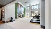This expansive fitness center offers a thoughtfully designed, single-floor layout that accommodates a full range of workout experiences. The space is divided into distinct functional zones, each with its own character and purpose. The main cardio area commands immediate attention with its soaring double-height ceilings, large floor-to-ceiling windows that flood the room with natural light, and views of lush outdoor surroundings. Ceiling fans provide airflow throughout, while a dramatic large-scale athletic mural and a vertical living green wall create a vibrant, energizing atmosphere. Spirit-brand treadmills, ellipticals, and cable machines are arranged to maximize open floor space. Transitioning deeper into the facility, a dedicated strength training zone features a comprehensive selection of selectorized machines and a dumbbell rack, complemented by floor-to-ceiling mirrors and a bold illuminated "SWEAT IT" sign that reinforces the motivational ambiance. Dark ceilings with recessed lighting contrast the lighter main area, lending this zone a focused, boutique-gym feel. Built-in shelving and cabinetry provide convenient storage. A wide, tile-floored corridor connects the zones and leads to the main entrance, lined with track lighting and accented by the recurring green wall motif. The overall design blends contemporary aesthetics with functional fitness infrastructure.