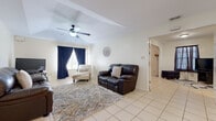 Located at 1602 S Airport Dr in Weslaco, Texas, this single-story home offers a practical and comfortable layout with consistent light tile flooring throughout. The living room is a standout space, featuring a vaulted tray ceiling with a ceiling fan, crisp white walls, and natural light filtering through navy-curtained windows. Elegant arched doorways connect the living area to the rest of the home, adding a subtle architectural detail. The kitchen is well-equipped with warm wood cabinetry, a mosaic tile backsplash, stainless steel refrigerator, and a peninsula that doubles as a breakfast bar. An adjacent dining area comfortably accommodates a dining table and chairs, creating a cohesive kitchen-dining space. The bedroom is a restful retreat with tile flooring, draping dark curtains for light control, and a rustic reclaimed wood headboard that adds character. The full bathroom features a wooden vanity with ample counter space and storage, a large mirror, and a tub-shower combination. A dedicated laundry room houses a full-size washer and dryer with overhead shelving for added storage convenience. Overall, the home presents a functional and well-maintained interior suited for comfortable everyday living.