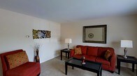 This two-floor residence offers a thoughtfully arranged living space designed for everyday comfort. The living room serves as a central gathering area, featuring wall-to-wall carpeting in a neutral tone that contributes to a warm, inviting atmosphere. Smooth, light-colored walls provide a clean backdrop throughout, complemented by standard-height ceilings that lend the room a comfortable sense of scale.

The living room is furnished with a rich, deep-red upholstered sofa accented with decorative throw pillows, paired with coordinating seating and a dark-finished coffee table that anchors the space. A matching end table holds a classic table lamp, providing ambient lighting. Decorative elements include an abstract metallic wall sculpture and a large framed mirror that enhances the sense of openness in the room. A tall decorative vase with branch arrangements adds a natural, artistic touch to one corner.

The overall ambiance of the living area is relaxed and neutral, with warm color accents creating visual interest against the subdued walls and flooring. The two-story layout offers additional living or sleeping space across both levels, providing functional versatility throughout the home.