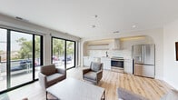 This contemporary single-floor space is defined by its clean lines, open-concept layout, and an abundance of natural light. Light hardwood flooring flows consistently throughout, complementing the crisp white walls and creating a bright, cohesive aesthetic.

The living area is a standout feature, lined with multiple floor-to-ceiling black-framed sliding glass doors that open directly onto a generous outdoor deck. The deck is furnished with comfortable seating and offers sweeping views of the surrounding cityscape and tree canopy, making it a seamless extension of the interior.

The kitchen is thoughtfully designed with sleek white cabinetry, a marble-look countertop, a white tile backsplash, and warm wood open shelving with integrated accent lighting. Stainless steel appliances — including a range, dishwasher, and refrigerator — complete the well-appointed culinary space, all framed by a distinctive arched architectural detail overhead.

Adjacent to the kitchen, the dining area features a large white-topped table illuminated by sculptural pendant lighting, with a sliding glass door providing direct deck access and views. A welcoming lounge area with a wall-mounted display screen and restroom facilities rounds out the interior, making this a highly functional and visually refined space suited for both everyday use and entertaining.