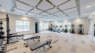 Located at 1840 Silas Deane Hwy in Rocky Hill, CT, this modern fitness center offers a generously sized, single-floor layout designed to accommodate a full range of workout activities. The space is divided into two distinct zones that flow seamlessly into one another, providing dedicated areas for both cardio and strength training.

The main cardio area features a striking coffered ceiling with geometric grid detailing and recessed lighting, lending the space a polished, contemporary aesthetic. Patterned carpet tiles in neutral tones cover the floor, complementing the light gray walls. Multiple large windows line the perimeter, filling the room with natural light and offering views of the surrounding area. Equipment includes treadmills, elliptical trainers, and stair climbers arranged in a row along the windows, alongside a wall-mounted flat-screen TV and full-length mirrors.

The adjoining strength and functional training zone boasts a lighter hardwood-style floor, a wall-mounted TV, TRX suspension trainers, a multi-use power rack, medicine balls, dumbbells, benches, rowing machines, and a Matrix stationary bike. A bold, artistic mural depicting athletic figures serves as a dynamic focal point on one wall, reinforcing the space's energetic atmosphere. The tray ceiling with layered lighting adds architectural interest to this section. Together, both areas create a comprehensive, well-appointed fitness facility.