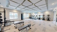 Located at 1840 Silas Deane Hwy in Rocky Hill, CT, this modern fitness center offers a generously sized, single-floor layout designed to accommodate a full range of workout activities. The space is divided into two distinct zones that flow seamlessly into one another, providing dedicated areas for both cardio and strength training.

The main cardio area features a striking coffered ceiling with geometric grid detailing and recessed lighting, lending the space a polished, contemporary aesthetic. Patterned carpet tiles in neutral tones cover the floor, complementing the light gray walls. Multiple large windows line the perimeter, filling the room with natural light and offering views of the surrounding area. Equipment includes treadmills, elliptical trainers, and stair climbers arranged in a row along the windows, alongside a wall-mounted flat-screen TV and full-length mirrors.

The adjoining strength and functional training zone boasts a lighter hardwood-style floor, a wall-mounted TV, TRX suspension trainers, a multi-use power rack, medicine balls, dumbbells, benches, rowing machines, and a Matrix stationary bike. A bold, artistic mural depicting athletic figures serves as a dynamic focal point on one wall, reinforcing the space's energetic atmosphere. The tray ceiling with layered lighting adds architectural interest to this section. Together, both areas create a comprehensive, well-appointed fitness facility.