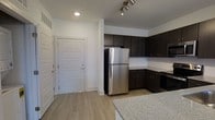 This single-floor space showcases a clean, modern design with a neutral color palette and thoughtful layout. The kitchen is a standout feature, equipped with dark espresso-toned cabinetry that provides ample storage, stainless steel appliances including a full-size refrigerator, and granite-look countertops that add a refined touch. A track lighting fixture overhead ensures the kitchen is well-lit and functional. The light wood-tone flooring runs throughout, creating a warm and cohesive aesthetic that complements the white walls and trim. The entry door, finished in crisp white with panel detailing, is positioned adjacent to the kitchen, creating a seamless transition into the living space. Recessed lighting in the ceiling contributes to a bright, contemporary atmosphere. The open-concept design between the kitchen and entry area maximizes the sense of space, making the layout feel efficient and practical. The overall ambiance is modern and move-in ready, with a neutral design that offers flexibility for personalization.