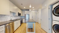 This single-floor residence showcases a thoughtfully designed open layout that seamlessly connects the kitchen and living areas. The kitchen is a standout feature, finished with flat-panel white cabinetry extending to the ceiling for generous storage capacity. Dark charcoal countertops provide a striking contrast against the crisp white surfaces, while a white subway tile backsplash adds texture and a clean aesthetic. A full suite of stainless steel appliances — including a refrigerator, electric range, built-in microwave, and dishwasher — equips the space for practical everyday use. A freestanding central island with a matching dark countertop and open lower shelf serves as both a workspace and a casual dining area, anchored by pendant lighting overhead. Light-toned hardwood flooring runs throughout the main living areas, contributing to the bright and airy atmosphere created by recessed lighting. The bedroom is carpeted in a neutral tone and benefits from a large window that invites natural light and views of the surrounding neighborhood. The bathroom features a glass-enclosed walk-in shower with large-format white wall tiles, mosaic tile flooring, and chrome fixtures, offering a clean, contemporary finish throughout.