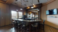 This pub is a single-floor venue thoughtfully designed to balance a social bar atmosphere with a relaxed dining environment. The space centers around a dark wood bar with a smooth countertop surface, fitted with pendant lighting overhead and a mirrored back bar that reflects the surrounding space, creating a sense of depth and openness. Overhead glass rack storage and built-in cabinetry add functional elegance to the bar area, which accommodates seating along multiple sides.

The main dining area extends generously from the bar, featuring warm hardwood flooring throughout that lends a classic, inviting character. Dining tables of varying sizes — including round tops and four-top configurations — are paired with ladder-back chairs and cushioned seating to suit a range of group sizes. Upholstered booth-style seating lines portions of the walls, offering additional comfort for guests.

Tall ceilings with exposed structural beams add architectural interest, while multiple windows fitted with wood-slat blinds line the walls, allowing natural light to fill the room. Pendant and track lighting supplements the natural illumination. Wall-mounted art and a flat-screen display contribute to the pub's character. A glass-pane exterior door provides easy access, reinforcing the open and approachable nature of this well-appointed establishment.