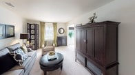 This thoughtfully designed single-floor residence offers a cohesive layout with multiple living spaces, two bedrooms, and two bathrooms. The living room is generously proportioned, featuring plush carpeting, neutral walls, and large windows dressed with elegant gold drapes that invite natural light. A round leather ottoman serves as a central focal point, complemented by a substantial dark wood armoire and a decorative folding screen. An adjacent sitting area provides a quiet retreat, accented by a charming window with a roman shade, a houndstooth-patterned chair, and a lush indoor palm. The kitchen is well-equipped with warm oak cabinetry, dark granite-style countertops, black appliances including a range, microwave, refrigerator, and dishwasher, and light tile flooring. The primary bedroom is a serene space with a ceiling fan, plush carpet, dark curtains, a tufted upholstered bed frame, and ample natural light. The second bedroom offers similar comfort with an upholstered headboard and coordinated décor. Both bathrooms feature tile flooring, single vanities with storage, and a tub-shower combination. The overall ambiance is warm, sophisticated, and cohesive throughout.