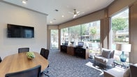 This single-story business center offers a thoughtfully designed workspace that balances functionality with a welcoming ambiance. The room is anchored by a workstation area along the far wall, where two desktop computer setups are arranged on a warm wood-toned desk, providing dedicated spaces for focused work or printing tasks. A ceiling fan overhead ensures comfortable air circulation throughout the space.

Large floor-to-ceiling windows span the exterior wall, flooding the interior with abundant natural light while offering expansive views of an outdoor pool area and lush landscaping beyond. A glass door provides direct access to the outdoor space, enhancing the open, connected feel of the room.

The patterned carpet in cool gray and blue tones adds texture underfoot and complements the neutral wall tones. A round wooden conference table with ergonomic task chairs occupies the left side of the space, making it well-suited for small meetings or collaborative work sessions. A wall-mounted flat-screen television is positioned nearby for presentations or media use. A comfortable lounge chair in the corner rounds out the space, offering a casual seating option for reading or relaxed work.