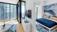 This contemporary single-floor residence showcases a clean, modern design defined by crisp white walls, light hardwood flooring, and an open-concept layout that maximizes both space and functionality. The living area flows seamlessly from the kitchen, creating an inviting, connected environment accented by curated artwork and bold blue décor elements that add visual interest against the neutral backdrop.

The kitchen is sleekly designed with flat-panel white cabinetry extending to the ceiling, a stainless steel gas range, and an undermount sink — offering a minimalist yet highly functional cooking space. Recessed lighting highlights the streamlined countertops and cabinetry.

The living and sleeping areas benefit from dramatic floor-to-ceiling windows framed in dark metal, flooding the space with abundant natural light and offering expansive, elevated views of the surrounding cityscape. The bedroom connects directly to the main living area, continuing the cohesive design language throughout.

The bathroom is a refined retreat featuring large-format marble-look wall tiles surrounding a full soaking tub with shower, complemented by a floating white double-vanity with an integrated countertop, backlit mirror, and textured penny-tile flooring — a sophisticated blend of texture and tone. The overall ambiance is polished, bright, and thoughtfully curated for modern urban living.