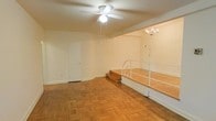 This single-floor space showcases a distinctive split-level layout that adds architectural interest and defined zoning within an open floor plan. The lower level is finished with classic parquet hardwood flooring in a warm honey tone, lending a timeless and elegant character to the space. A white ceiling fan with a built-in light fixture is centrally positioned overhead, providing both ventilation and ambient lighting.

A set of wooden steps with a white metal railing leads up to a raised platform area, which is finished with matching wood tones and illuminated by a decorative chandelier, creating a naturally distinguished secondary zone ideal for dining or a reading nook. The raised section also features a wall-mounted light fixture, adding layered lighting options.

The walls are painted in a clean, neutral white throughout, contributing to a bright and airy atmosphere. White baseboards frame the perimeter cleanly, and a solid interior door provides access to an additional room or closet. The overall ambiance is calm and versatile, offering a blank canvas well-suited to a variety of furnishing and design styles.