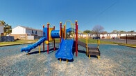 Located at 181 Derrer Rd in Columbus, Ohio, this outdoor playground area serves as a dedicated recreational space within a residential community. The centerpiece of the space is a vibrant, multi-component play structure finished in bold blue, red, and yellow. The structure includes multiple slides — featuring both a wide blue slide and a secondary slide — along with a tunnel, climbing panels, and connecting platforms, offering a variety of play experiences for children of different ages and abilities.

The play area is surfaced with a generous spread of loose gravel, providing impact-absorbing ground cover across the entire footprint of the space. The layout offers ample open area surrounding the equipment, giving children room to move freely. A dedicated lighting pole is positioned within the space, extending usability into evening hours.

The playground is enclosed on one side by a wooden privacy fence and is flanked by neatly maintained grassy lawns, with the surrounding residential buildings visible in the background. The open-sky setting allows for abundant natural light throughout the day. The overall environment is clean, well-maintained, and thoughtfully designed to provide a safe and engaging outdoor amenity for the surrounding community.