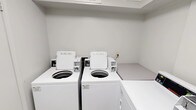 This laundry facility is a single-floor, self-contained room designed for practical, efficient use. The space features white walls and light tile flooring, creating a clean and bright environment. The room is equipped with multiple commercial-grade Speed Queen top-load washing machines and front-load dryers, all finished in white and arranged along the walls to maximize the available floor space. A built-in folding counter adjacent to the machines provides a convenient surface for sorting and folding laundry. The machines are managed through a CSC payment system, which supports mobile app-based payments, with instructional signage posted prominently on the wall for user guidance. Overhead shelving above the machines offers additional storage or staging space. The room is accessed through a solid white door fitted with a keyed entry lock, ensuring secure access. A wall-mounted light switch controls the interior lighting. The overall design of the space is utilitarian and straightforward, prioritizing function and ease of use. Signage reminds users to remove clothing promptly upon cycle completion, reflecting a well-maintained and community-oriented shared facility.