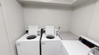 This laundry facility is a single-floor, self-contained room designed for practical, efficient use. The space features white walls and light tile flooring, creating a clean and bright environment. The room is equipped with multiple commercial-grade Speed Queen top-load washing machines and front-load dryers, all finished in white and arranged along the walls to maximize the available floor space. A built-in folding counter adjacent to the machines provides a convenient surface for sorting and folding laundry. The machines are managed through a CSC payment system, which supports mobile app-based payments, with instructional signage posted prominently on the wall for user guidance. Overhead shelving above the machines offers additional storage or staging space. The room is accessed through a solid white door fitted with a keyed entry lock, ensuring secure access. A wall-mounted light switch controls the interior lighting. The overall design of the space is utilitarian and straightforward, prioritizing function and ease of use. Signage reminds users to remove clothing promptly upon cycle completion, reflecting a well-maintained and community-oriented shared facility.