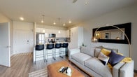 This single-floor residence showcases a thoughtfully designed open-concept layout that seamlessly integrates the kitchen, dining, and living areas. The kitchen is a standout feature, equipped with high-gloss white cabinetry, dark countertops, stainless steel appliances—including a refrigerator, microwave, and gas range—and a large island with pendant lighting overhead, providing both additional prep space and casual seating. A smart home panel is conveniently positioned near the kitchen entrance.

The living area flows naturally from the kitchen, featuring wood-style flooring, a ceiling fan, and large windows alongside a sliding glass door that opens to a balcony, filling the space with natural light and outdoor views. Recessed lighting complements the warm, contemporary ambiance throughout.

The bedroom offers plush carpeting, a window with outdoor views, and access to a generously sized walk-in closet fitted with double hanging rods and shelving. The bathroom features wood-style flooring, a tub-shower combination with tile surround, a white vanity with dark countertop, and a well-lit mirror. Neutral tones and modern finishes unify the space with a clean, cohesive aesthetic.