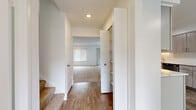 This well-maintained two-floor residential unit offers a bright, neutral interior with a clean, modern aesthetic throughout. The galley-style kitchen is a standout feature, equipped with stainless steel appliances including a range, dishwasher, and refrigerator. Light wood-toned cabinetry with sleek bar-pull hardware lines both walls, complemented by white quartz countertops and a classic subway tile backsplash. Wood-look flooring runs through the kitchen and hallway areas, adding warmth and durability. A built-in floor-to-ceiling storage cabinet in the main hallway provides convenient organizational space, while recessed lighting illuminates the corridor leading between floors. The spacious living area features plush wall-to-wall carpeting, large sliding glass doors dressed with vertical blinds for natural light and outdoor access, and a stylish chandelier overhead. Additional windows with horizontal blinds further brighten the space. Carpeted bedrooms are generously sized with neutral white walls and ceiling vents for climate control. The bathroom includes a wide vanity with light wood cabinetry, white countertop, a large mirror, and a combined tub and shower enclosure. A staircase with carpeted treads connects the two floors, completing this functional and move-in-ready home.