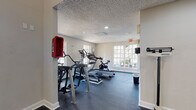 This fitness center in Jacksonville, Florida, is designed to support a well-rounded exercise routine within a clean, functional environment. The space is organized into distinct workout zones, allowing users to move efficiently between cardio and strength training areas.

The cardio section features a selection of machines, including a treadmill, elliptical, and stationary cycle, positioned along windowed walls that allow natural light to filter in and provide views of the surrounding outdoor landscape. A textured ceiling with recessed lighting adds to the bright, open feel of this area.

The strength training section is notably spacious, with soaring ceilings and large multi-pane windows that flood the room with daylight. Wall-mounted mirrors enhance the sense of openness and allow users to monitor their form. A multi-station cable machine paired with an adjustable bench offers a versatile range of resistance exercises. A dedicated free weight area includes a full rack of dumbbells and a standing scale for tracking progress.

Throughout the facility, dark rubberized flooring provides durability and comfort underfoot. Safety features, including a mounted fire extinguisher and clearly marked exit signage, are visible throughout. The neutral wall tones and clean layout contribute to a focused, welcoming workout environment.