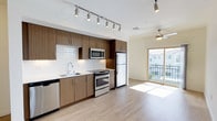 This single-floor space showcases a thoughtfully designed open-concept layout that seamlessly connects the kitchen and living area. The contemporary kitchen is outfitted with rich, medium-toned wood-grain cabinetry that extends to the ceiling, providing generous storage. A clean white subway tile backsplash contrasts beautifully against the cabinetry, while a full suite of stainless steel appliances — including a French door refrigerator, electric range, built-in microwave, and dishwasher — adds a polished, modern touch. The undermount sink with a sleek arc faucet is positioned beneath a bank of upper cabinets, maximizing workspace efficiency.

Underfoot, light-toned hardwood-style flooring runs throughout, creating a bright and cohesive feel. Overhead, a track lighting system in the kitchen offers focused illumination, while a ceiling fan in the living area promotes airflow and comfort. Large sliding glass doors at the far end of the living space flood the interior with abundant natural light and open onto a private balcony, offering views of the surrounding community and outdoor scenery. The overall ambiance is clean, airy, and contemporary.
