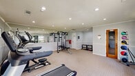 Located at 120 S Cranford Rd in Goose Creek, South Carolina, this dedicated exercise room offers a comprehensive fitness facility within a compact, efficiently utilized single-floor layout. The room features light gray walls accented by a white chair rail that runs the perimeter, crown molding, and neutral patterned carpeting that lends a clean, polished aesthetic throughout.

Recessed lighting provides bright, even illumination across the entire space, complemented by natural light from several windows dressed with simple curtain panels. A large wall mirror spans one side of the room, visually expanding the space and allowing users to monitor their form during workouts.

The exercise room is outfitted with multiple cardio machines, including treadmills and an elliptical trainer, as well as a multi-function cable resistance machine ideal for a full-body strength workout. A dedicated free weights area features a tiered dumbbell rack with a range of weights, while a medicine ball tower offers additional functional training options. A ballet barre is also present, accommodating stretching and balance exercises.

Finishing details include a textured ceiling, a fire safety station, a sanitizer dispenser, and access to a restroom, ensuring both safety and convenience for users. Overall, this fitness room provides a well-rounded and functional environment suited for a variety of workout routines.