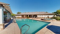 Located at 9270 Eagle Ranch Rd NW in Albuquerque, New Mexico, this single-level outdoor amenity area is designed to take full advantage of the sunny high-desert climate. The centerpiece of the space is a generously sized swimming pool with clear turquoise water, surrounded by a broad concrete deck furnished with multiple lounge chairs and shaded umbrellas — ideal for relaxing and sunbathing. A raised spa with stone surround sits adjacent to the main pool, offering a dedicated area for hydrotherapy and leisure. A handsome wood-beamed pergola provides a shaded outdoor living area outfitted with comfortable seating, perfect for socializing or unwinding out of the sun. A community building with stone-accented stucco exterior lines one side of the pool deck, housing additional amenities and facilities. The grounds are thoughtfully landscaped with mature shade trees, manicured hedges, and water-conscious xeriscape featuring decorative gravel and native plantings. Gated entry with iron fencing ensures a secure and private environment. The overall ambiance reflects the classic Southwestern architectural aesthetic, blending natural stone, earth tones, and lush greenery against the backdrop of Albuquerque's characteristic bright blue skies.